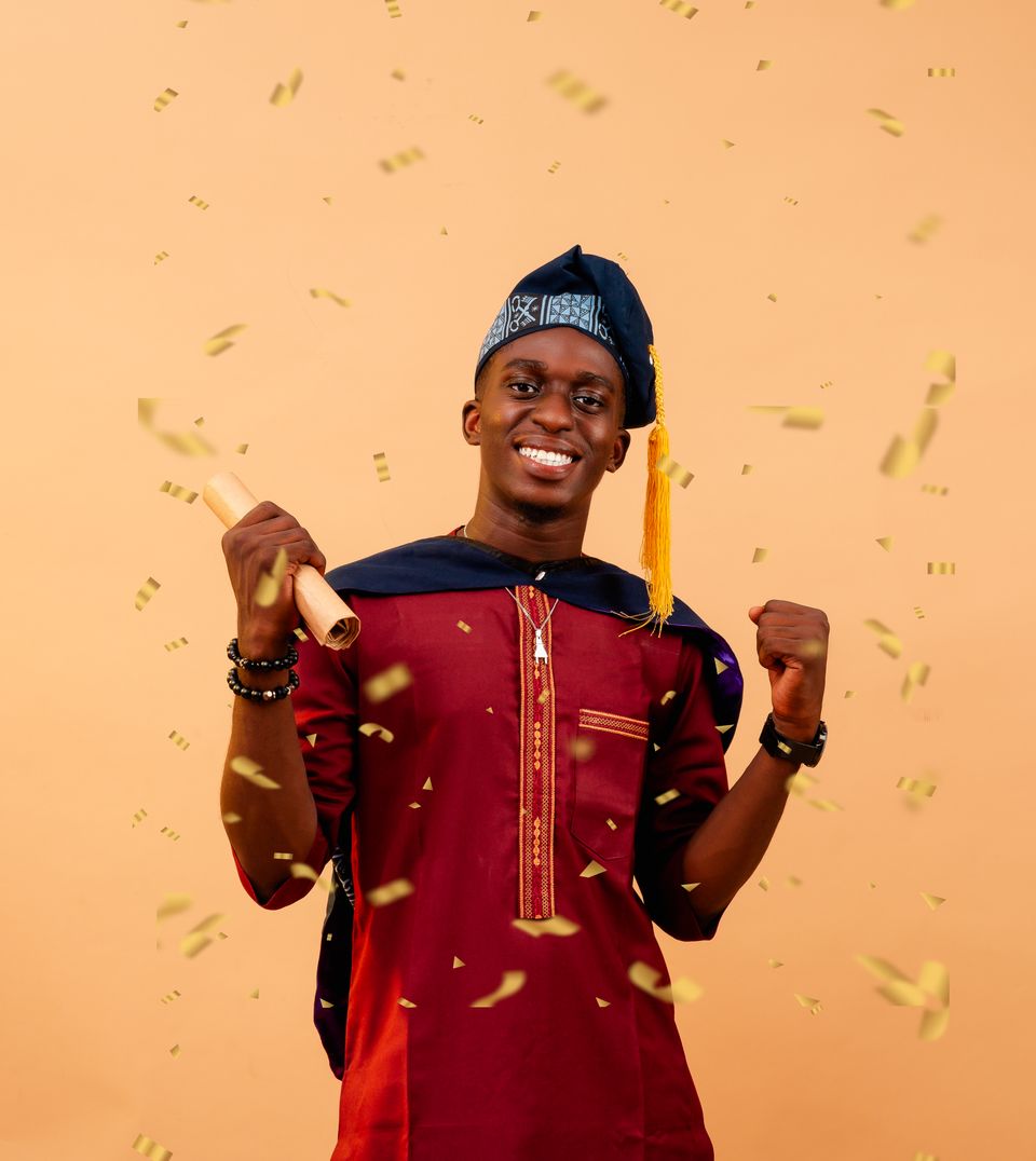 A beaming graduate in traditional attire raising his fist amid gold confetti